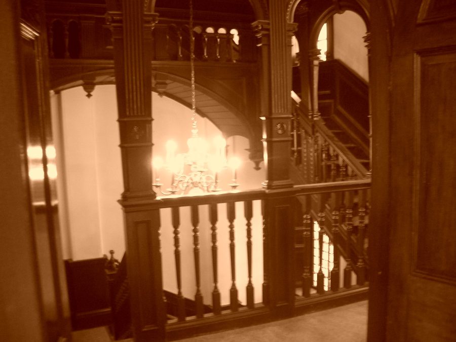 The main staircase: fluted posts, carved arches, a galleried landing and a candle-style chandelier hanging through the stairwell.